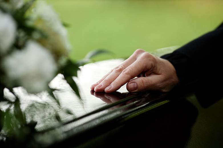 A hand rests on a casket at a funeral, suggesting loss and mourning.